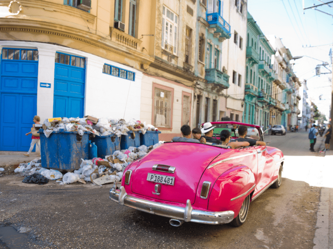 Carro con turistas pasa por un basurero en una esquina de una calle.
