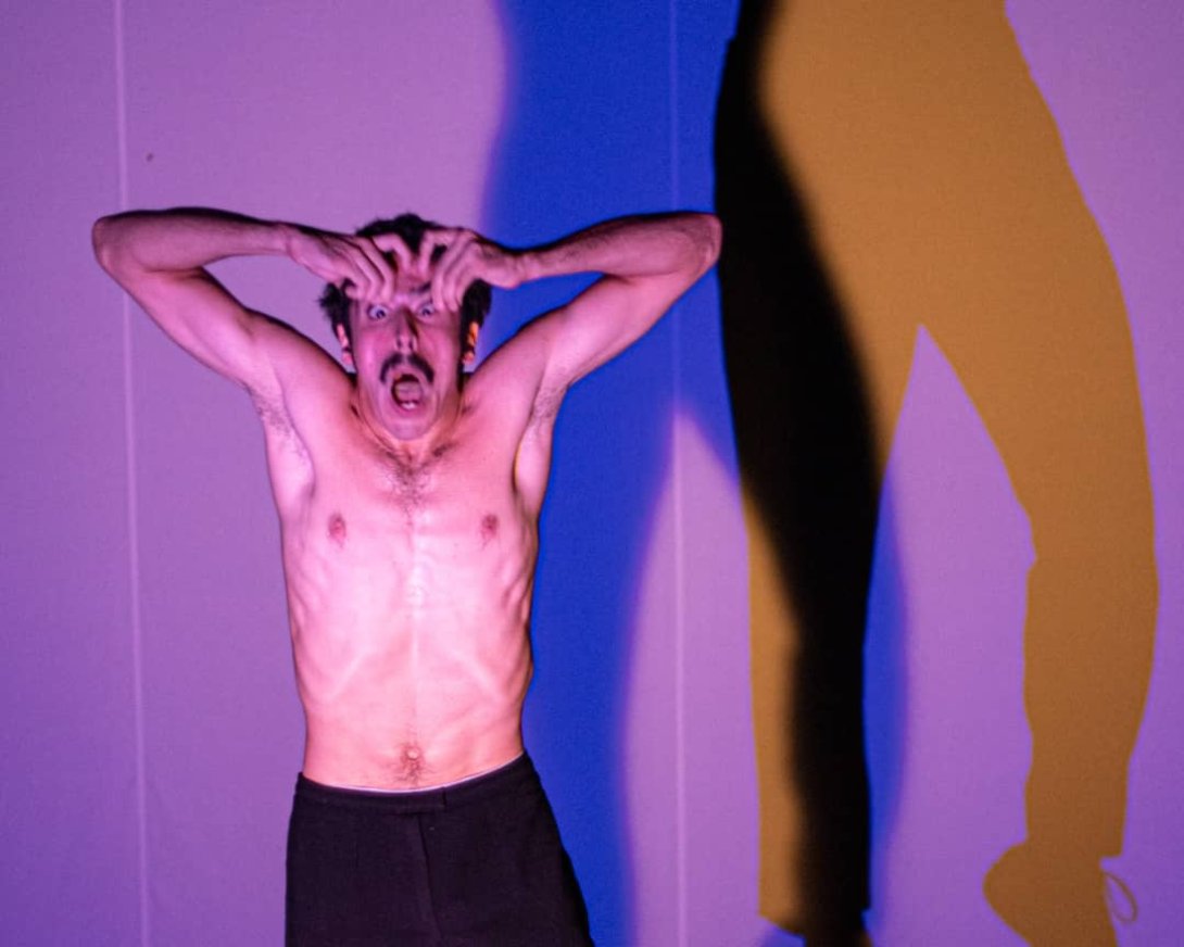 César Saavedra durante la puesta en escena de "Normalización": con el torso desnudo se lleva las manos a la cabeza, al fondo sombras de colores.