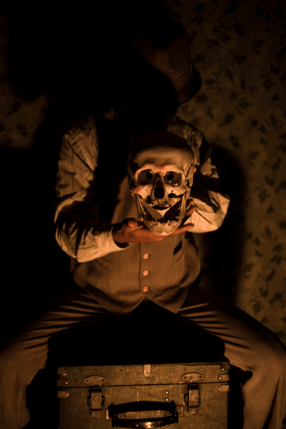 Actor sentado sobre una maleta en la oscuridad muestra una máscara entre sus manos.