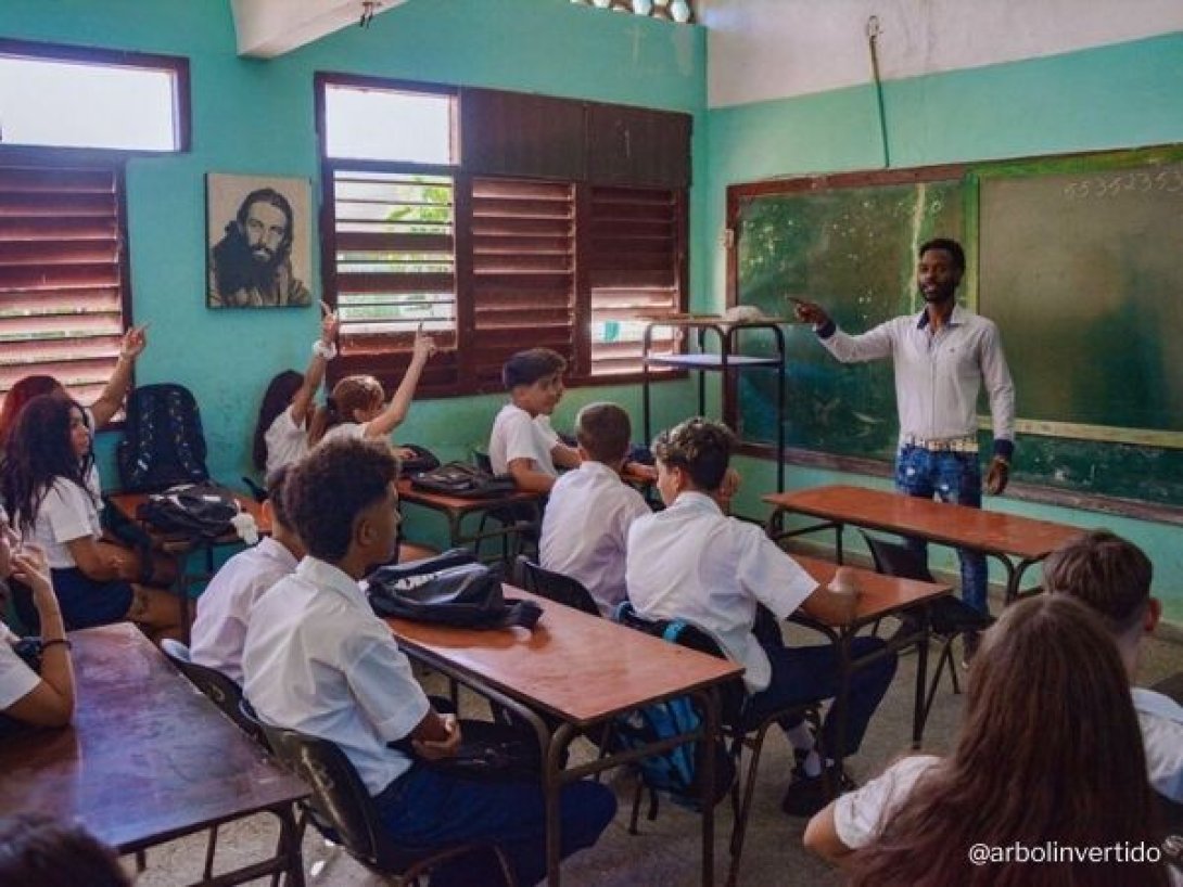 Aula en Cuba con estudiantes uniformados y un maestro frente a un pizarrón deteriorado.