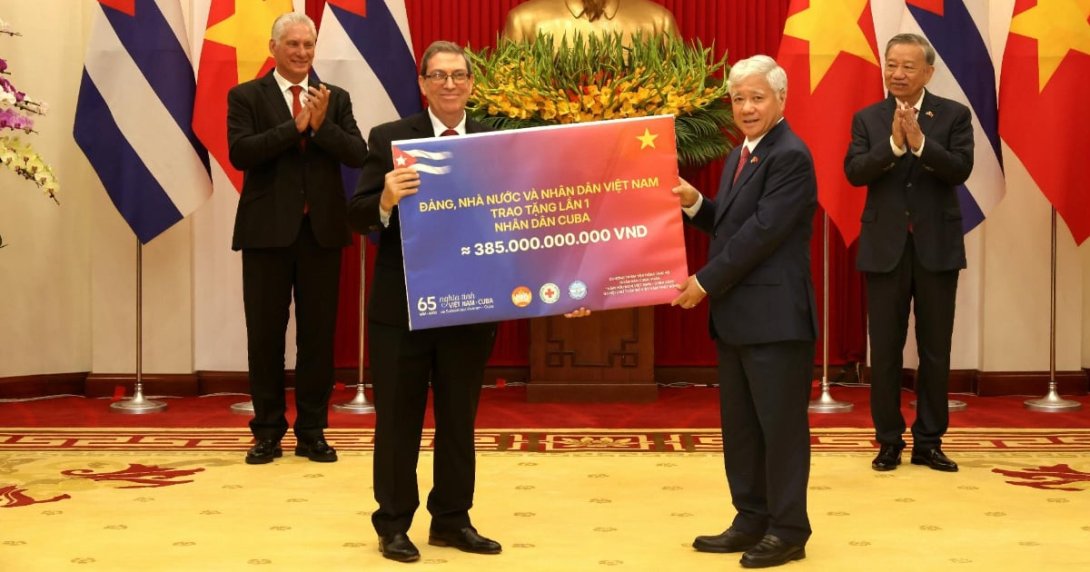 El canciller cubano Bruno Rodríguez y un funcionario vietnamita muestran un cheque simbólico de 15 millones de dólares donados a Cuba, con Díaz-Canel aplaudiendo detrás, en Hanói.