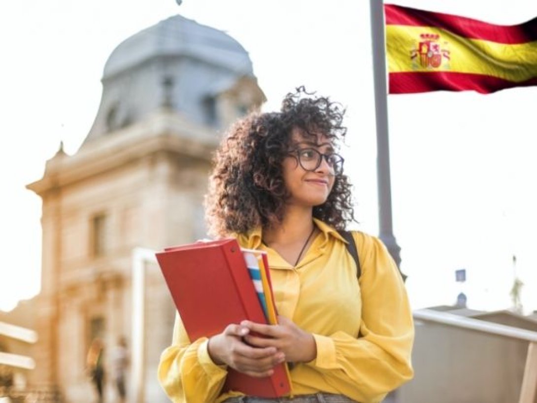 Estudiante joven con libros en la mano frente a un edificio histórico en España, junto a una bandera española.