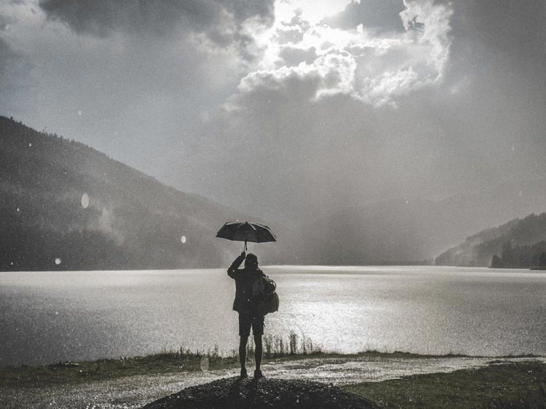 Mujer con sombrilla de frente a un lago mientras llueve.