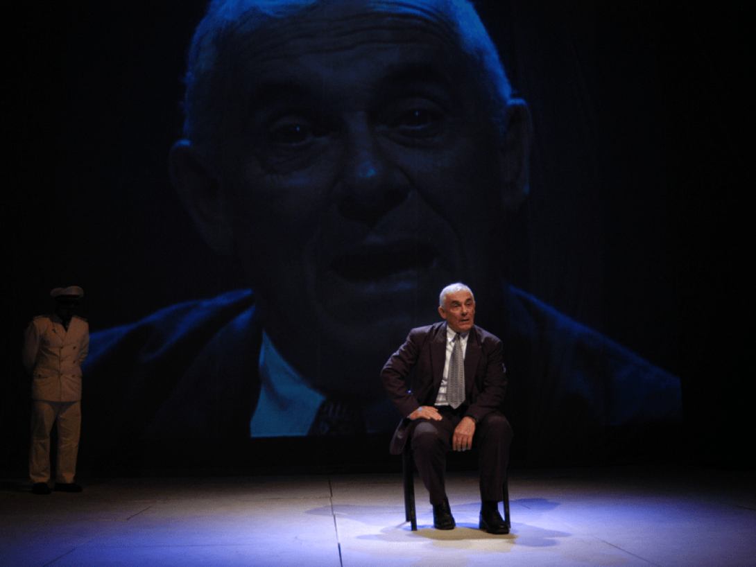 Pancho García en escena sentado sobre una silla, en la pantalla detrás se proyecto su rostro.