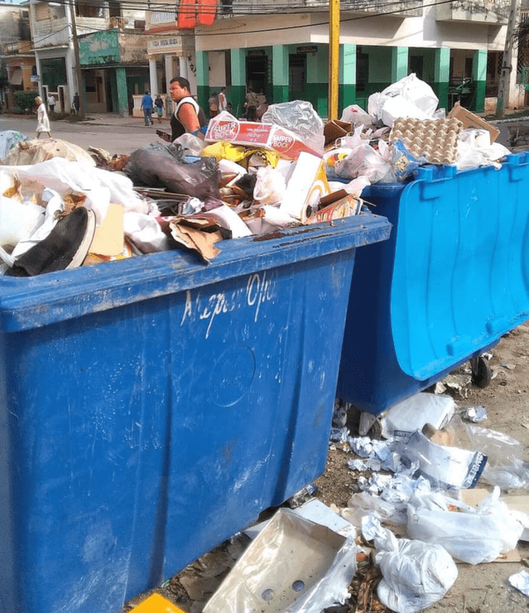 Dos contenedores azules de basura desbordan desperdicios hacia la calle. 