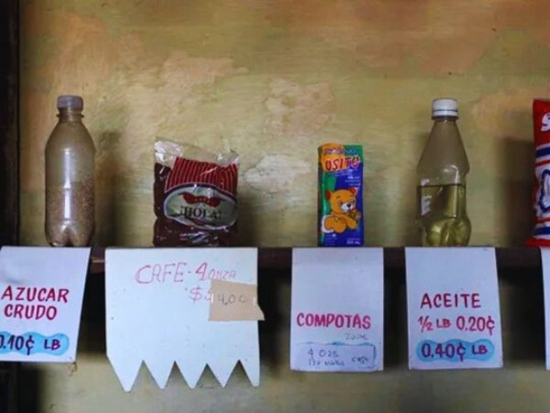 Estantes de una bodega cubana con productos racionados como azúcar, café, compotas y aceite, acompañados de carteles con precios subsidiados.