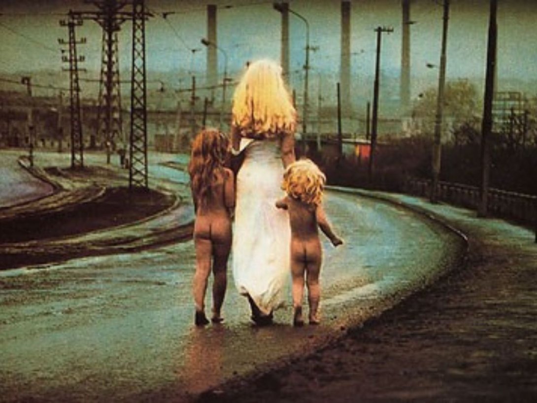 una mujer rubia con un vestido blanco camina hacia el horizonte tomando de la mano a dos niños desnudos