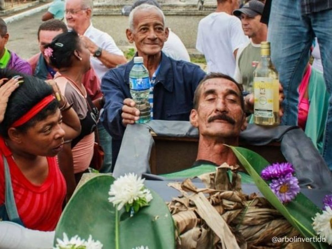 Hombre simula estar muerto en un ataúd durante el Velorio de Pachencho en Santiago de las Vegas, rodeado de vecinos con ron y flores.