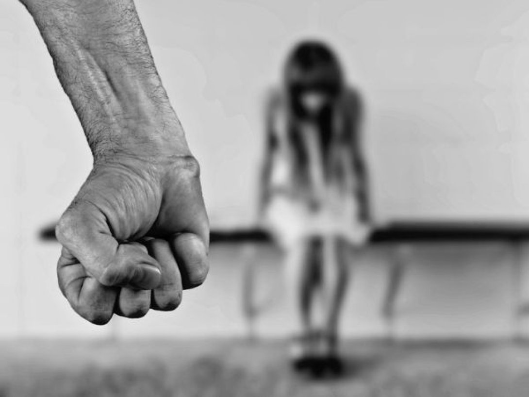 Mano de un hombre en actitud violenta frente a una mujer sentada con gesto de miedo y desamparo.