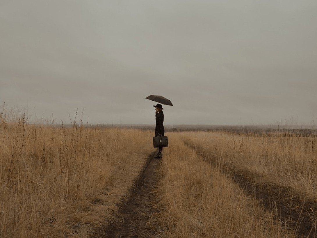 Una mujer con sombrilla y maleta en mano camina por un campo. 