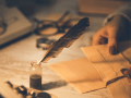 Manos femeninas sostienen papel para escribir, al lado la pluma y la tinta.