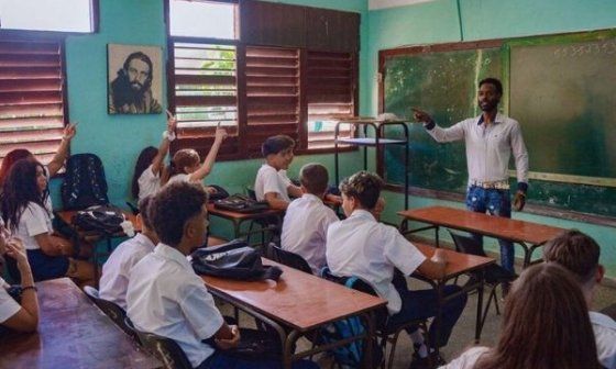 Aula en Cuba con estudiantes uniformados y un maestro frente a un pizarrón deteriorado.