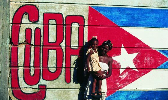 Mujer cargando una niña frente a una pared con la bandera cubana