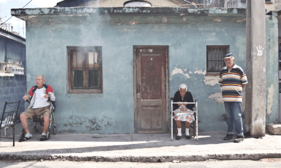 Ancianos esperan sentados en la calle mientras fumigan sus casas, Cuba. Sale humo por la ventana de una casa.