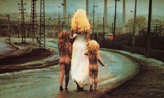 una mujer rubia con un vestido blanco camina hacia el horizonte tomando de la mano a dos niños desnudos