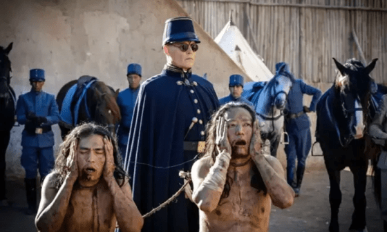 Dos personas arrodilladas y amarradas se llevan las manos a la cabeza en señal de dolor o desconcierto. De pie, otros hombre vestidos de uniforme y con caballos los escoltan.